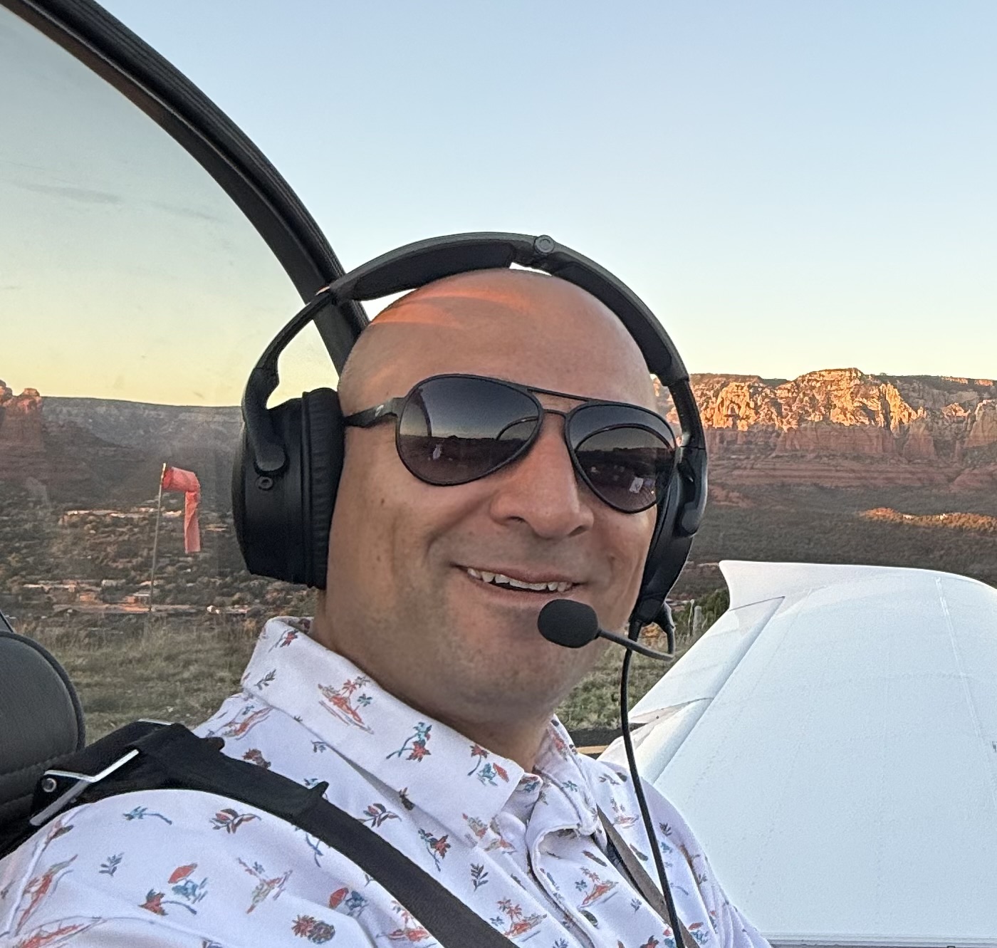 Rich Wiens in the cockpit wearing a headset and aviator sunglasses, with red rock cliffs behind him at sunset